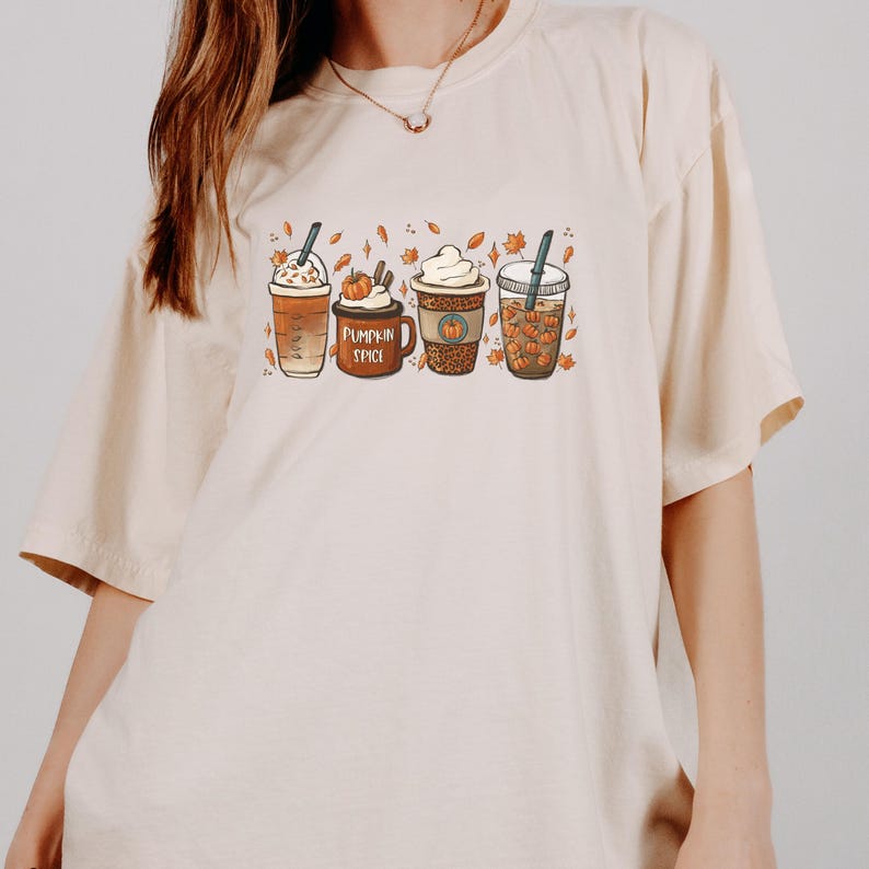 May include: A beige t-shirt with a graphic of four different fall-themed coffee drinks. The drinks are decorated with fall leaves and pumpkins. The text "Pumpkin Spice" is printed on the shirt.