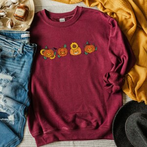 May include: A maroon sweatshirt featuring a line of four smiling jack-o'-lantern pumpkins with sunflowers and vines. The pumpkins are orange with black eyes, noses, and mouths.