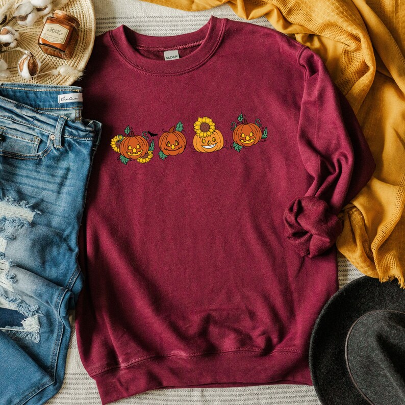May include: A maroon sweatshirt featuring a line of four smiling jack-o'-lantern pumpkins with sunflowers and vines. The pumpkins are orange with black eyes, noses, and mouths.