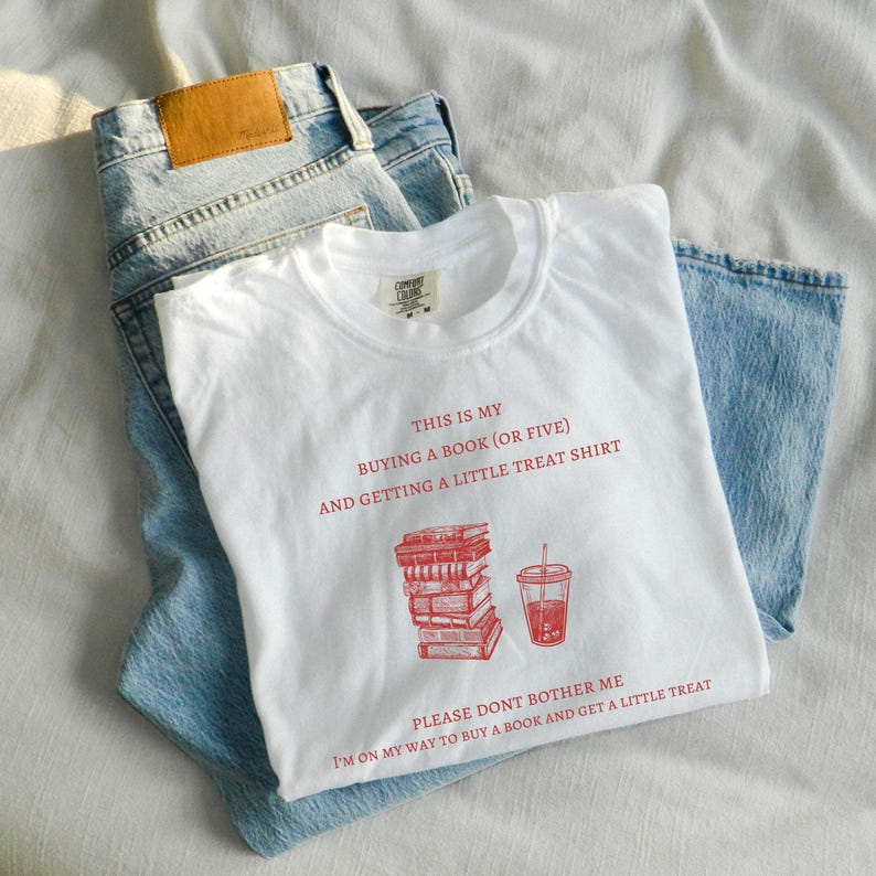 May include: White t-shirt with red text and graphics, including a stack of books and a drink. The shirt reads "THIS IS MY BUYING A BOOK (OR FIVE) AND GETTING A LITTLE TREAT SHIRT" and "PLEASE DONT BOTHER ME I'M ON MY WAY TO BUY A BOOK AND GET A LITTLE TREAT". Light blue jeans are also in the image.