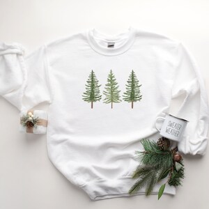 May include: White sweatshirt with a graphic of three green watercolor pine trees. The sweatshirt is laying flat on a white surface with a white mug that says "Sweater Weather" and a sprig of pine.