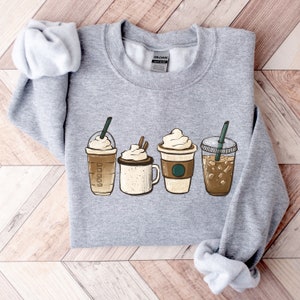 Könnte beinhalten: Ein hellgraues Sweatshirt mit vier Abbildungen von Kaffeegetränken mit Schlagsahne und Strohhalmen. Die Kaffeetassen sind braun, beige und transparent. Das Sweatshirt besteht aus einem weichen Material, ideal für Kaffeeliebhaber.