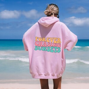 May include: Pink hoodie with the text "FOREVER CHASING SUNSETS" in a retro font with colorful letters. The hoodie is worn by a person standing on a beach.