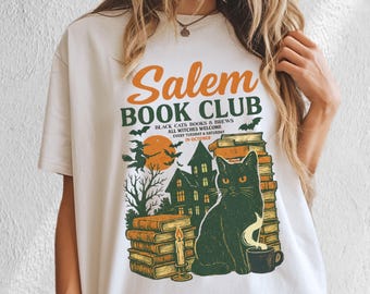 Puede incluir: Camiseta color crema con un diseño gráfico que presenta el texto "Salem Book Club". El diseño incluye un gato negro, pilas de libros, una casa embrujada, murciélagos y el texto "Black Cats, Books & Brews, All Witches Welcome".