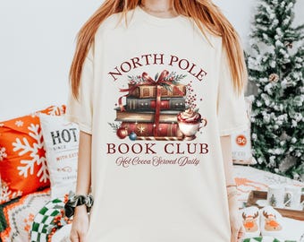 May include: A white t-shirt with a red and green design that says "North Pole Book Club" with a stack of books, a cup of hot cocoa, and the text "Hot Cocoa Served Daily".
