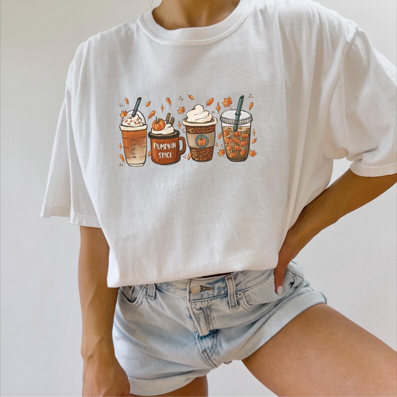 May include: White t-shirt with a graphic of four fall-themed coffee drinks. The drinks are decorated with fall leaves and pumpkins. The text "Pumpkin Spice" is on the cup in the center.