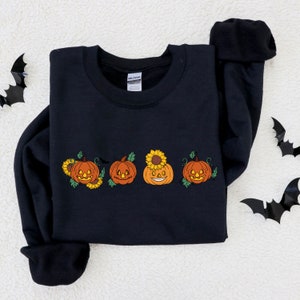 May include: Black sweatshirt featuring a design of four jack-o'-lantern pumpkins with sunflowers on top. The pumpkins have different facial expressions.