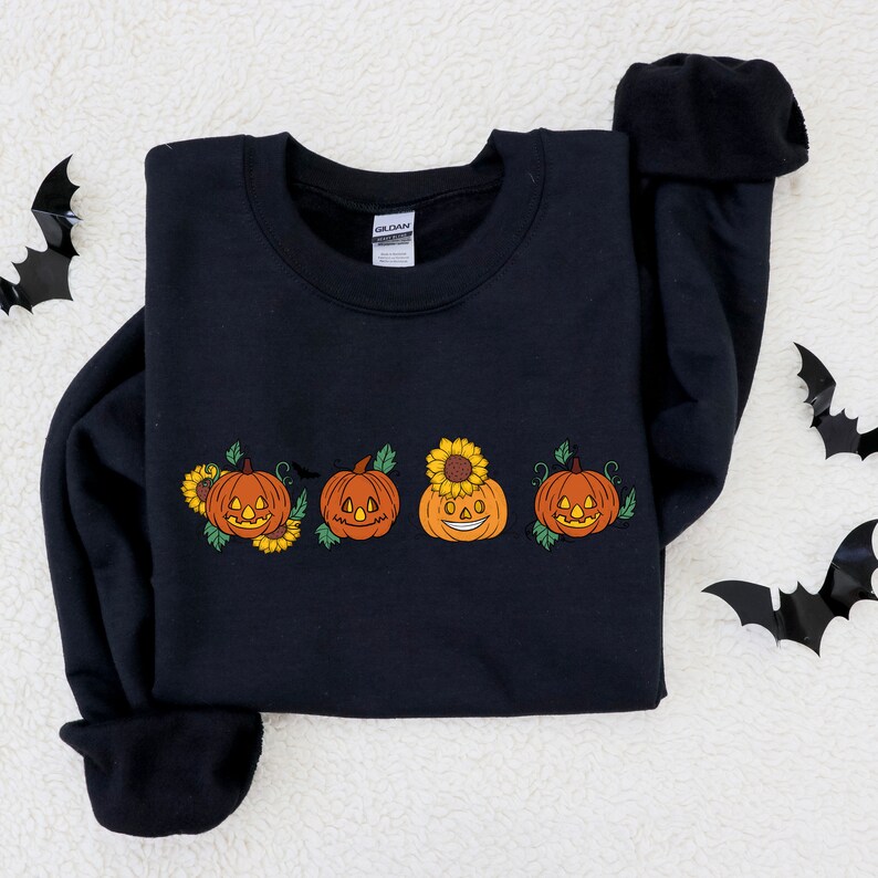 May include: Black sweatshirt featuring a design of four jack-o'-lantern pumpkins with sunflowers on top. The pumpkins have different facial expressions.