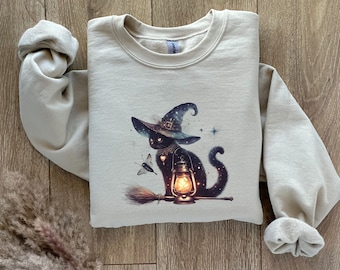 Puede incluir: Una sudadera beige con un gato negro que lleva un sombrero de bruja y sostiene una linterna. El gato está rodeado de estrellas y una mariposa. La sudadera tiene cuello redondo y mangas largas.