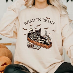 May include: A beige sweatshirt with the text "Read in Peace" printed on the front. The graphic features a skeleton lying in a coffin reading a book. There are bats and candles surrounding the coffin.