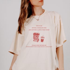 May include: Cream-colored t-shirt with red text and illustrations. The text reads "THIS IS MY BUYING A BOOK (OR FIVE) AND GETTING A LITTLE TREAT SHIRT" and "PLEASE DONT BOTHER ME I'M ON MY WAY TO BUY A BOOK AND GET A LITTLE TREAT".