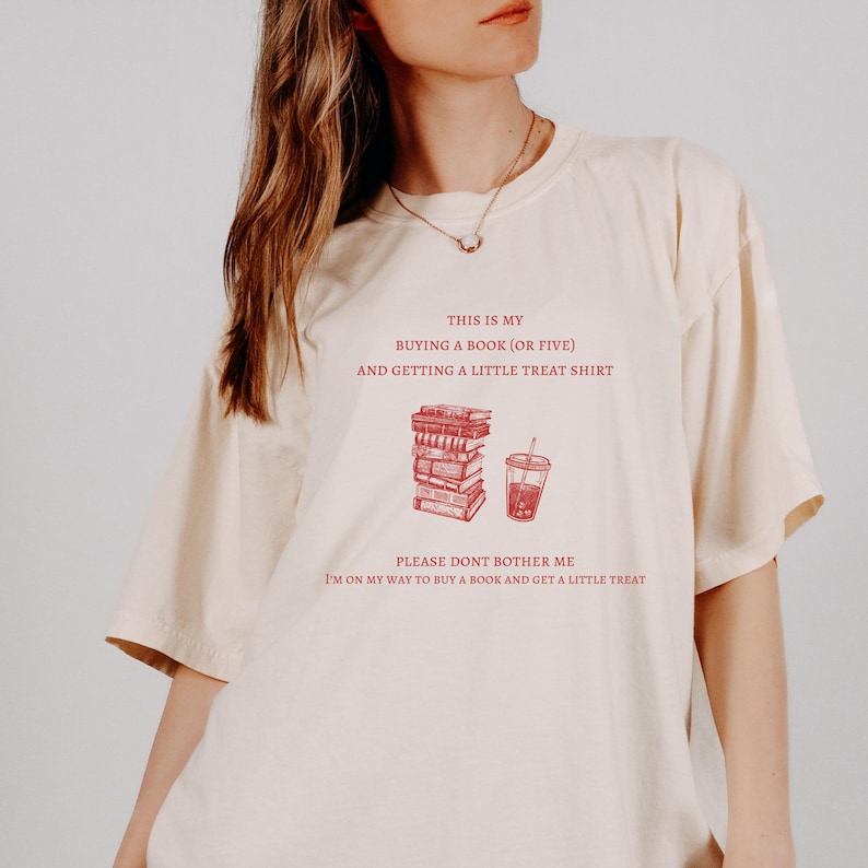 May include: Cream-colored t-shirt with red text and illustrations. The text reads "THIS IS MY BUYING A BOOK (OR FIVE) AND GETTING A LITTLE TREAT SHIRT" and "PLEASE DONT BOTHER ME I'M ON MY WAY TO BUY A BOOK AND GET A LITTLE TREAT".