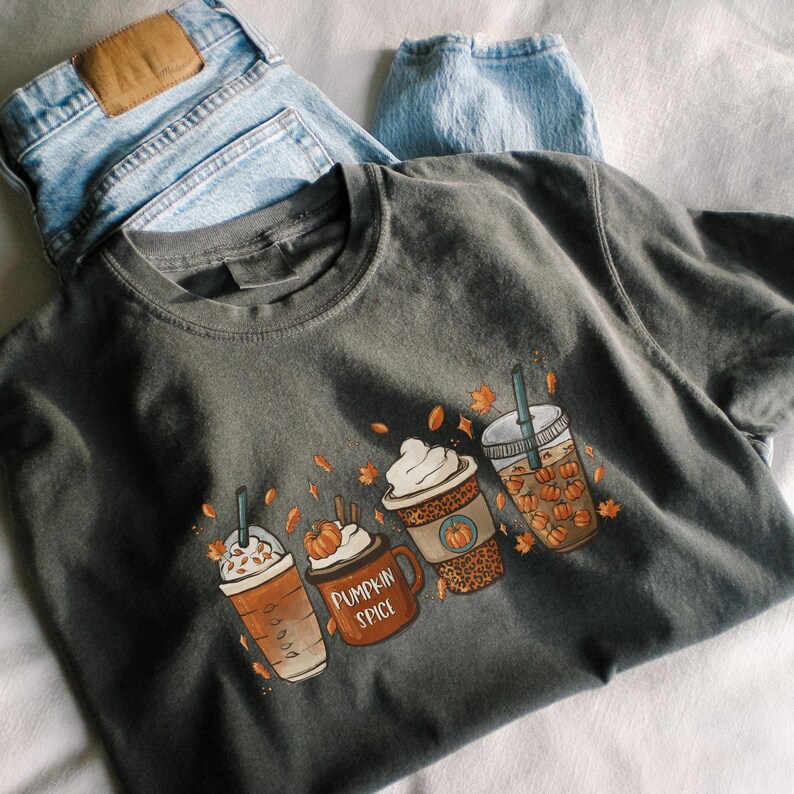 May include: A dark gray t-shirt with a graphic of four fall-themed coffee drinks. The drinks are decorated with whipped cream, pumpkin spice, and fall leaves. The text "Pumpkin Spice" is visible on one of the drinks.