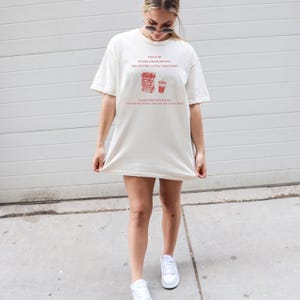 May include: Cream-colored t-shirt with red text that reads "THIS IS MY BUYING A BOOK (OR FIVE!) AND GETTING A LITTLE TREAT SHIRT." The shirt also has a graphic of books and a drink. The text continues, "PLEASE DON'T BOTHER ME. I'M ON MY WAY TO BUY A BOOK AND GET A LITTLE TREAT!"