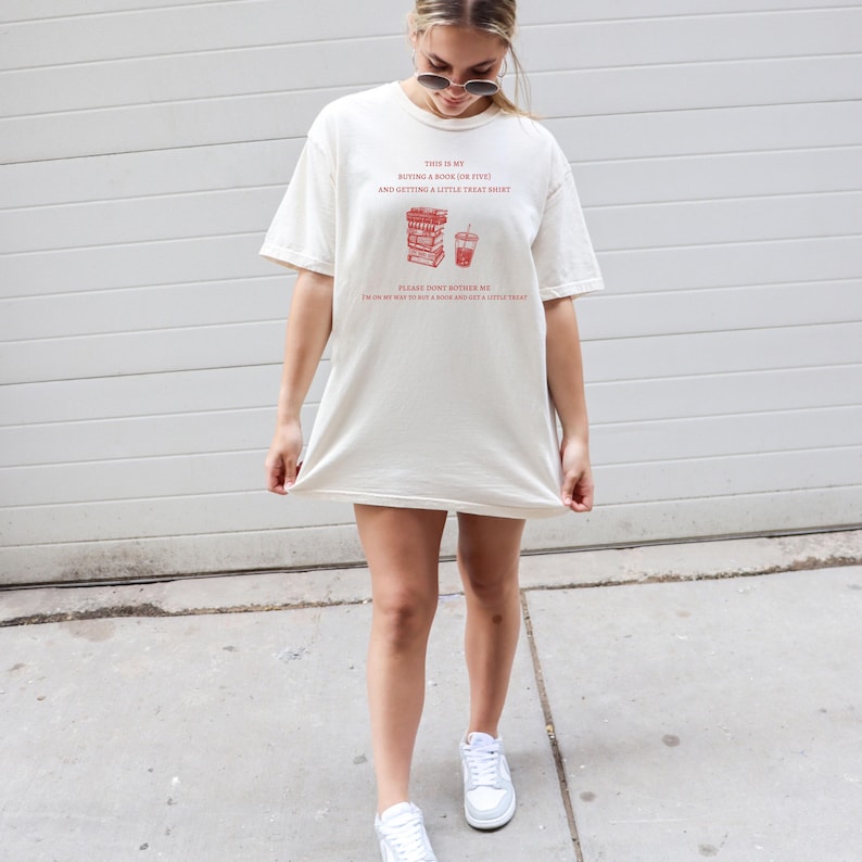 May include: Cream-colored t-shirt with red text that reads "THIS IS MY BUYING A BOOK (OR FIVE!) AND GETTING A LITTLE TREAT SHIRT." The shirt also has a graphic of books and a drink. The text continues, "PLEASE DON'T BOTHER ME. I'M ON MY WAY TO BUY A BOOK AND GET A LITTLE TREAT!"