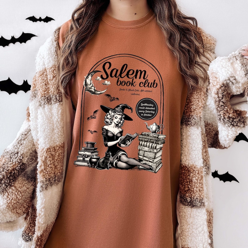 Puede incluir: Una sudadera marr&oacute;n con un dise&ntilde;o gr&aacute;fico de una bruja leyendo un libro frente a una pila de libros. El gr&aacute;fico tambi&eacute;n incluye una luna creciente, murci&eacute;lagos y el texto "Salem Book Club".