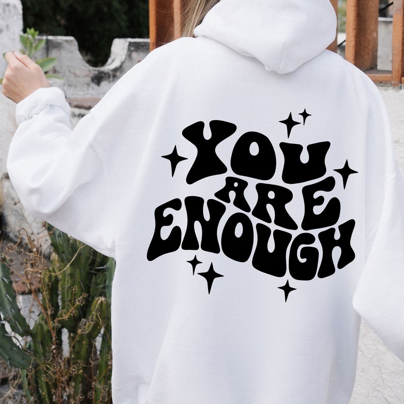 Puede incluir: Sudadera con capucha blanca con texto negro que dice "YOU ARE ENOUGH" en una fuente retro con estrellas alrededor del texto.