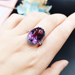 Pode incluir: Anel de ametista com uma grande gema roxa de corte oval. A pedra preciosa é rodeada por um halo de pequenas pedras transparentes e realçada com pedras de corte baguette. Montagem prateada.