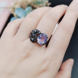 Peut inclure: Bague noire ornée d'une grosse pierre ovale violet clair. La bague présente un motif floral avec trois accents en forme de rose. La bague est portée sur une main.