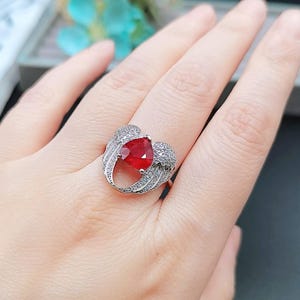 Könnte beinhalten: Ein silberner Ring mit einem roten herzförmigen Edelstein, der von klaren Steinen umgeben ist. Der Ring hat ein Design mit zwei Flügeln auf jeder Seite des Herzens.