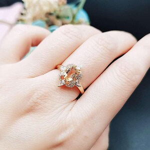 Pode incluir: Um anel de ouro com uma grande pedra preciosa ovalada cercada por pedras preciosas menores e transparentes. O anel está em um dedo.