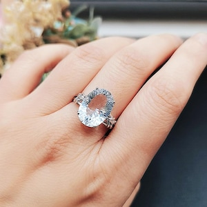 Pode incluir: Um anel de prata com uma pedra preciosa oval azul clara. O anel tem um design detalhado em forma de folha na banda. A pedra preciosa é o ponto focal, capturando a luz e criando um efeito brilhante. É uma joia.