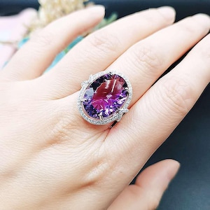 Pode incluir: Um anel de prata com uma grande ametista ovalada cercada por pequenas pedras preciosas transparentes.