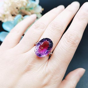 Może przedstawiać: Pierścionek z ametystem z dużym, owalnym kamieniem o fioletowo-czerwonym gradiencie kolorów. Pierścionek ma złoty pierścień z małymi kamieniami akcentującymi. Pierścionek jest prezentowany na dłoni.