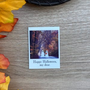 Könnte beinhalten: Ein rechteckiger Aufkleber mit einem Foto von zwei weißen Geistern, die sich auf einem Pfad durch einen herbstlichen Wald mit orangefarbenen und gelben Bäumen an den Händen halten. Unter dem Bild steht der Text "Happy Halloween, my dear." in schwarzer Serifenschrift.