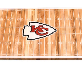May include: A wooden game table with a Kansas City Chiefs logo and a football field design. The table is painted with white lines and numbers, and the logo is red and white.