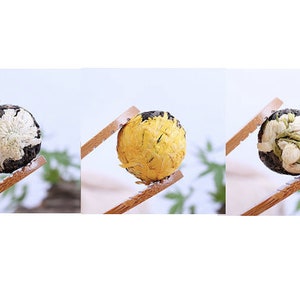 May include: Three tea balls, each with a different flower design. The first tea ball is white with a black center, the second is yellow, and the third is green with white flowers. The tea balls are held by wooden chopsticks.
