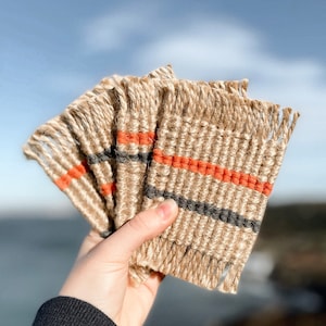 Könnte beinhalten: Set aus vier gewebten Untersetzern mit einer natürlichen Jutebasis und schwarzen und orangefarbenen Streifen. Die Untersetzer haben einen Fransenrand.