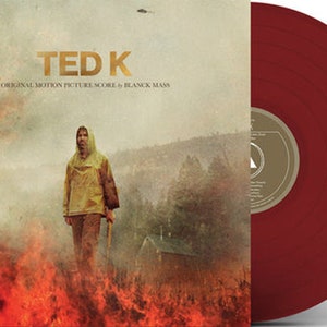 May include: A red vinyl record with a gold label. The record cover features a man in a yellow raincoat walking through a field of fire. The text on the cover reads "TED K ORIGINAL MOTION PICTURE SCORE by BLANCK MASS".
