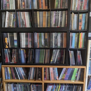 May include: A collection of vinyl records stored in black and wooden shelves. The records are arranged vertically, with their spines facing outward, displaying a variety of colors and designs. The shelves are filled with a diverse selection of music albums.