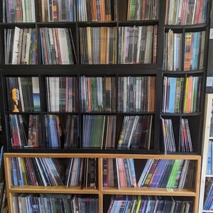 May include: A large collection of vinyl records displayed on black and wooden shelves. The records are stored vertically, with their spines visible, showcasing a range of colours and designs. The shelves are filled with a diverse selection of music albums.