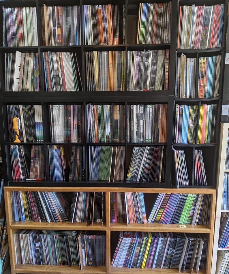 May include: A large collection of vinyl records displayed on black and wooden shelves. The records are stored vertically, with their spines visible, showcasing a range of colours and designs. The shelves are filled with a diverse selection of music albums.