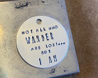 May include: A silver metal round tag with the text "Not all who wander are lost... but I am" in black lettering.