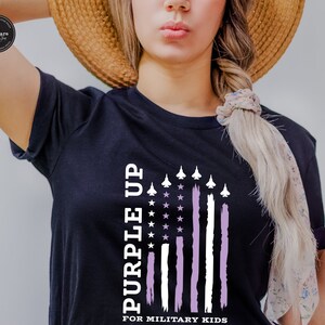 May include: A black t-shirt featuring a purple and white American flag design. The flag has white stars and white stripes with a purple stripe on the right side. The text "Purple Up" is written vertically on the left side of the flag and the text "For Military Kids" is written below the flag.