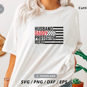 Op de afbeelding: Wit T-shirt met een zwarte en rode afbeelding met de tekst "Husband Daddy Protector Hero" met een klein Amerikaans vlagontwerp. De afbeelding is gecentreerd op het T-shirt.
