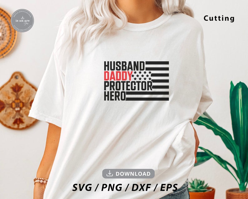 Op de afbeelding: Wit T-shirt met een zwarte en rode afbeelding met de tekst "Husband Daddy Protector Hero" met een klein Amerikaans vlagontwerp. De afbeelding is gecentreerd op het T-shirt.