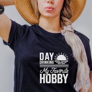 Puede incluir: Camiseta negra con texto blanco que dice "Day Drinking My Favorite Hobby" con un gr&aacute;fico de puesta de sol.