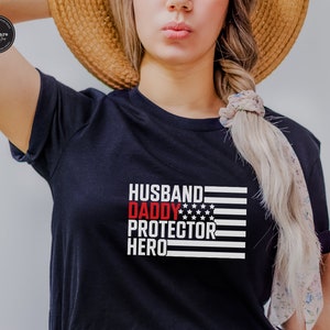 Op de afbeelding: Zwart T-shirt met een rood, wit en blauw Amerikaans vlagontwerp. De tekst "Husband Daddy Protector Hero" is in wit op de vlag gedrukt.