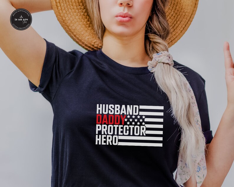 Op de afbeelding: Zwart T-shirt met een rood, wit en blauw Amerikaans vlagontwerp. De tekst "Husband Daddy Protector Hero" is in wit op de vlag gedrukt.