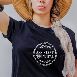 May include: A navy blue t-shirt with the words "Assistant Principal" in a white circular design. The design includes the words "Warm, Compassionate, Caring, Loving, Kind, Loyal, Reliable, Fun, Dedicated" around the central text.