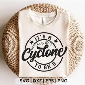 May include: Cream-colored t-shirt with a black circular design that reads "It's a great day to be a Cyclone." The design includes stars and the word "Cyclone" in a bold font. The shirt is displayed on a woven surface.