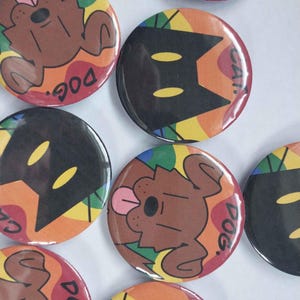 May include: A collection of round buttons with colorful designs. Some buttons feature a brown dog illustration with the word "DOG" in red. Other buttons have a black cat illustration with the word "CAT" in red. The background has a rainbow design.