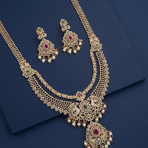 May include: A gold-toned necklace and earring set featuring intricate filigree patterns, clear crystals, teardrop-shaped pink stones, oval green stones, and dangling white pearls. The necklace includes a large peacock-themed pendant.