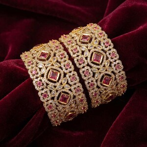 Könnte beinhalten: Zwei breite goldfarbene Armreifen mit einem komplizierten Filigranmuster, das mit kleinen klaren Kristallen und größeren quadratischen rosa Steinen besetzt ist. Das Metall zeigt ein geometrisches Muster mit einem zentralen quadratischen Stein.