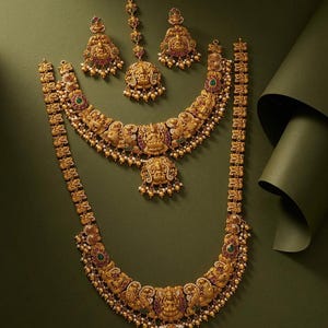 May include: A gold-toned jewelry set featuring two necklaces, a pair of earrings, and a maang tikka. Each piece is adorned with intricate deity motifs, red and green stones, and small white pearl-like beads. The text SS Traditions is visible.