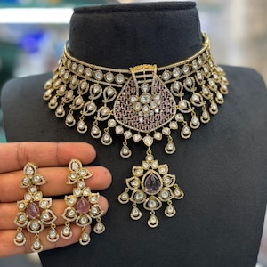 May include: A gold-toned necklace and matching earring set featuring intricate filigree metalwork, embedded clear stones, and deep red teardrop-shaped accents. The necklace includes a central purple stone cluster and dangling elements.